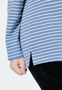 Blå randig långärmad topp med vita ränder, med sidoflikar vid fållen och mjukt bomullsmaterial. Svart ribbad tyg nedanför toppen.