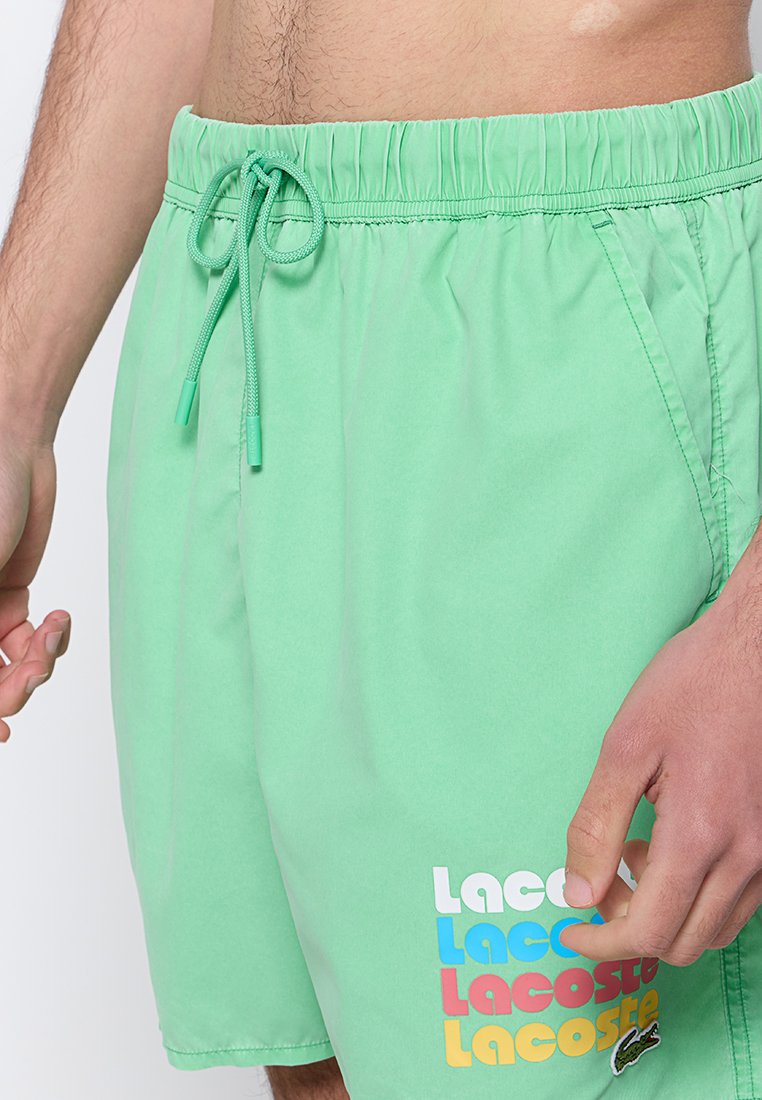 Jasnozielone szorty kąpielowe z elastycznym paskiem w talii i sznurkiem do regulacji. Z przodu na dole znajduje się wielokolorowe logo "Lacoste". Gładka tekstura.