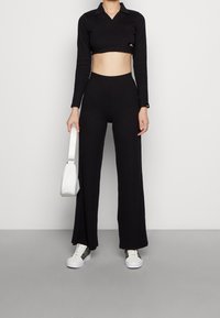 Schwarzes, kurzes langärmliges Oberteil mit Kragen, kombiniert mit hochtaillierten, weiten schwarzen Hosen. Das Model hält eine kleine weiße Handtasche.