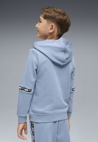 Ragazzo con capelli corti castani che indossa un felpa con cappuccio azzurro chiaro e pantaloni abbinati con strisce nere del logo Puma, che guarda verso sinistra, in piedi su uno sfondo grigio.