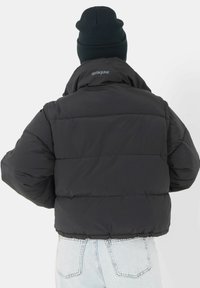 Zwarte pufferjas met een boxy vorm, voorzien van horizontale baffles, een hoge kraag en subtiele branding op de achterkant. Te dragen met lichtblauwe jeans.