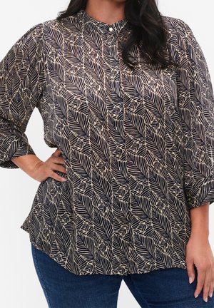 Frau, die eine langärmelige Bluse mit schwarzen und beigen Blattmustern und blaue Jeans trägt, mit der Hand in der Hüfte, vor einem schlichten Hintergrund stehend.