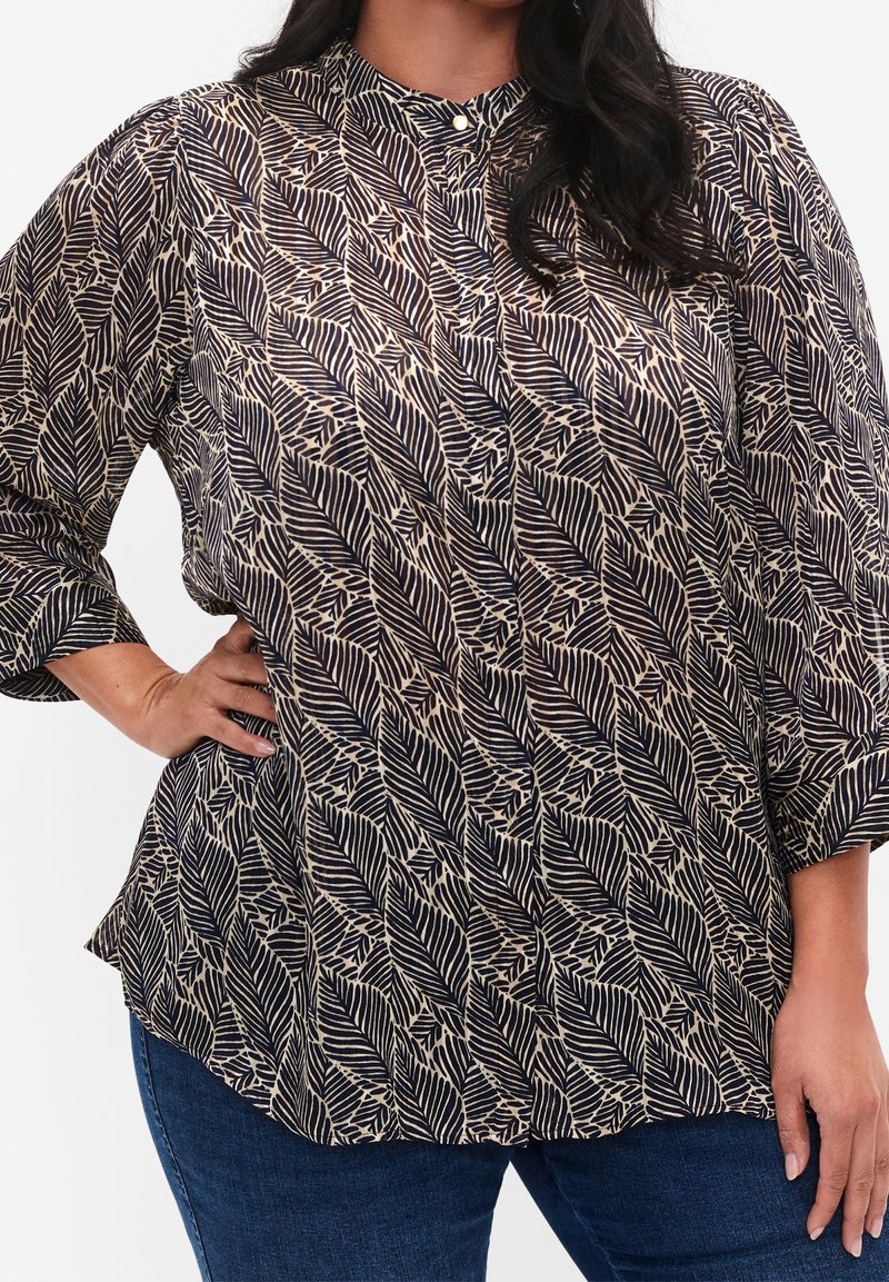 Femme portant un chemisier à manches longues avec des motifs de feuilles noirs et beiges et un jean bleu, main sur la hanche, debout devant un fond uni.
