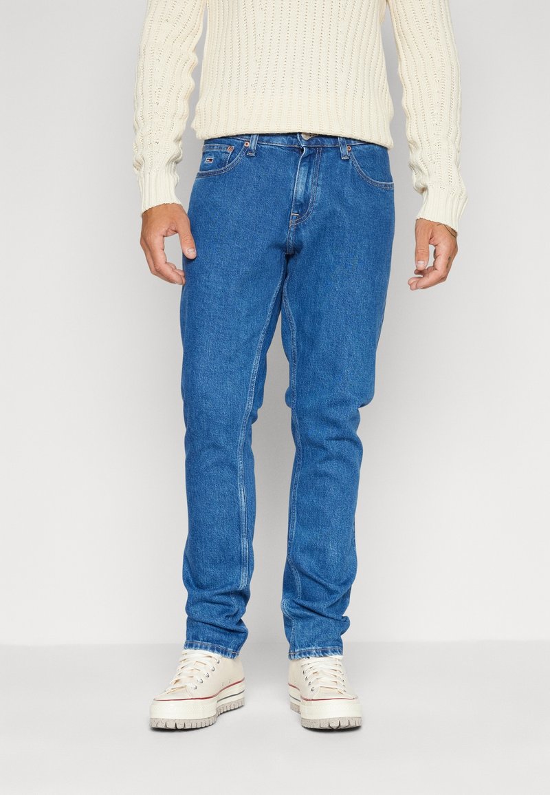 Modré džínové rifle s úzkým střihem, pět kapes a zipem na předním díle. Nositelná s krémovým pleteným svetrem a bílými teniskami.