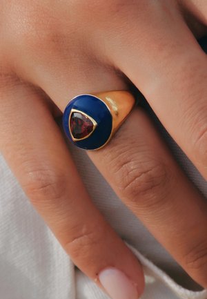 Bague en or avec un dôme en émail bleu, agrémentée d'une gemme rouge triangulaire au centre, portée sur une main au teint naturel et avec un léger vernis à ongles.