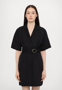 Rochie neagră cu curea, cu mâneci scurte, având un guler în formă de V și un detaliu cu cataramă metalică rasucită la talie. Textură fină a materialului.