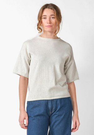 Junge Frau mit welligem braunem Haar trägt ein hellbeiges kurzärmeliges Oberteil und blaue Jeans, steht vor weißem Hintergrund.