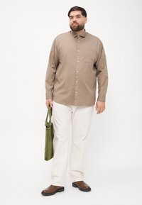 Hellbraunes Hemd mit Knöpfen, Kragen und Brusttasche, kombiniert mit cremefarbenen Hosen, hält eine grüne Tasche. Braune Schuhe.