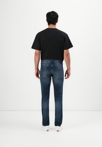 Mees, kes kannab pleekinud siniseid teksaseid, musta T-särki ja valgeid spordijalatsid, seisab seljaga lihtsalt valge tausta poole.