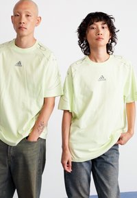 Světle zelené oversized trička s krátkým rukávem a kulatým výstřihem, zdobené jemným logem a abstraktními liniovými detaily.