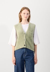 Groene, fluffy vest met een V-halsontwerp, voorzien van zes knopen en een gekruiste, hoekige zoom. Gepaard met losse denim broeken.
