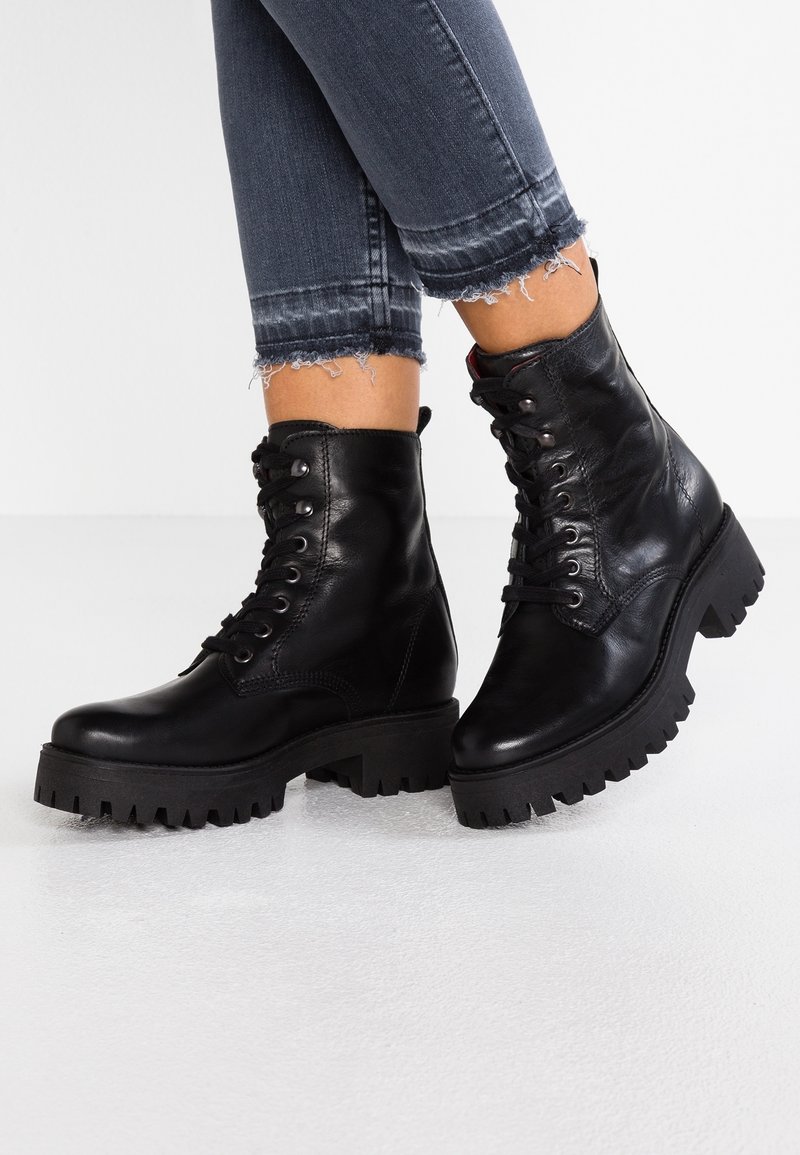 Černé kožené kotníkové boty na šněrování s masivní, strukturovanou podrážkou a polstrovaným límcem. Nosené s šedými džíny s ohrnutým lemem a roztrhanými okraji.