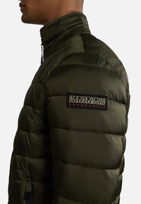 Giacca a vento verde oliva con colletto alto, texture trapuntata e zip laterale. Presenta un patch in tessuto con logo sulla spalla sinistra.