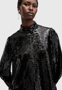 Top nero a maniche lunghe realizzato in paillettes lucide, con collo alto e spalle a sbuffo, creando una superficie riflettente e strutturata.