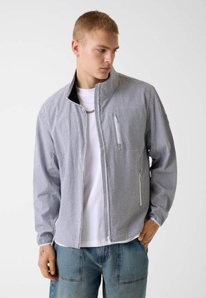 Giovane uomo che indossa una giacca con cerniera a quadri bianco e neri sopra una maglietta bianca e jeans blu, che guarda in basso verso sinistra.