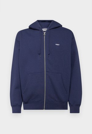 Mornarska zip-up dukserica s kapuljačom na vezivanje, prednjim džepovima, rebrastim manžetama i malim bijelim logotipom na lijevoj strani prsiju. Materijal je mješavina pamuka.