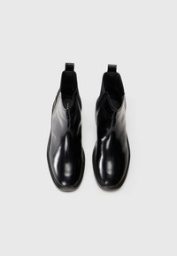 Un par de botines de cuero negro brillante, tipo slip-on, con lengüetas de tirón y punteras redondeadas cuadradas sobre un fondo blanco.