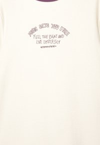 Witte langemouwen shirt met een paarse ronde hals. Het tekstontwerp bevat "MUZIEK MAKT JE VRIJ" en coördinaten eronder. Zachte textuur.
