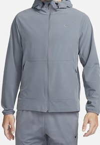 Grijze zip-up hoodie gemaakt van lichtgewicht materiaal. Voorzien van elastische manchetten, twee vo pockets en een subtiel logo op de borst.