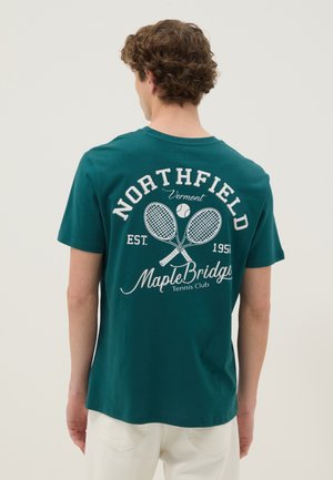 Homme vu de dos portant un t-shirt bleu sarcelle avec des raquettes de tennis blanches, une balle et le texte « Northfield Vermont Maple Bridge Tennis Club » dans le dos.