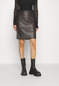 Femme portant une jupe en cuir brun foncé, un haut noir à manches longues avec des manches transparentes, et des bottes à plateforme noires épaisses, se tenant devant un fond blanc.