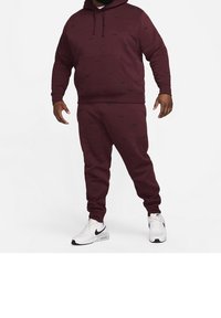 Burgundy hoodie with a front pocket and black pattern, paired with matching tapered pants. Complemented by white sneakers.