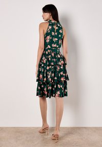 Robe florale verte avec un col halter et des volants superposés; présente des fleurs roses, une longueur courte et un détail de dos découpé. Portée avec des talons beiges.