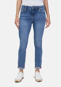 Jeans en denim bleu à coupe ajustée, présentant cinq poches, une fermeture à bouton classique et des ourlets roulés avec des accents rouges.