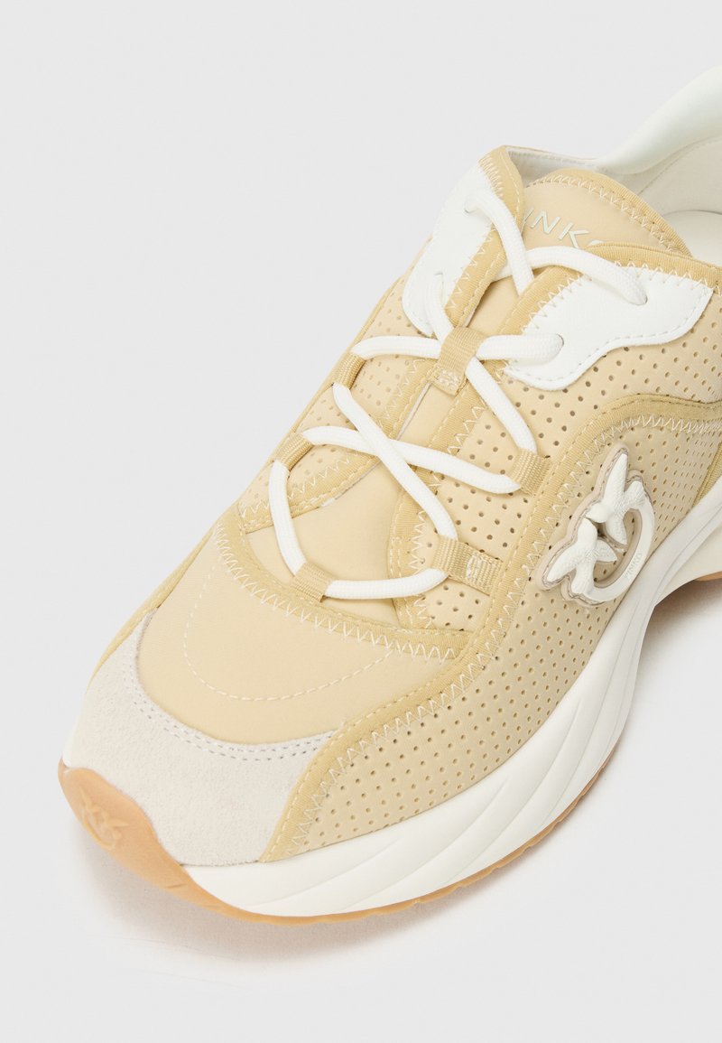 Chaussure de sport beige avec dessus en synthétique perforé, lacets blancs et accents en suède. Bout arrondi et semelle en caoutchouc épaisse. Détail de logo sur le côté.