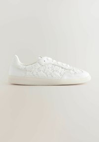Sneaker bianchi con sovrapposizione in pizzo floreale, materiale texturizzato, punta rotonda, suola piatta e dettagli lisci in contrasto. Design classico con lacci.