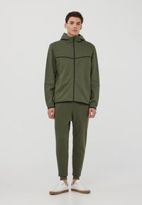 Felpa con cappuccio verde oliva, con zip frontale, dettagli neri e pantaloni coordinati, dotata di polsini a costine e una vestibilità comoda.