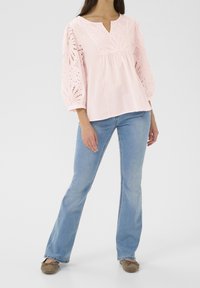 Ljusrosa blus med broderad ögla på trekvartsärmar, v-ringning, tillsammans med blå utsvängda jeans och bruna platta skor.