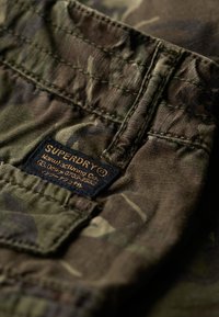 Tissu à motif camouflage avec des teintes de vert et de marron. Une étiquette noire avec un texte doré indique "SUPERDRY" cousue dans le matériau.
