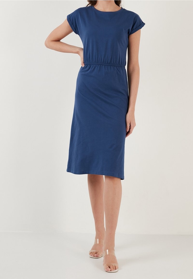 Robe bleu avec des manches courtes et une ceinture élastique, présentant un ourlet au niveau du genou et une texture douce et lisse. Sandales transparentes visibles.