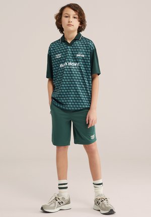 Fiatal személy zsebre dugott kézzel zöld mintás inget, hozzá illő rövidnadrágot, csíkos zoknit és szürke tornacipőt visel, egyszerű háttér előtt áll.