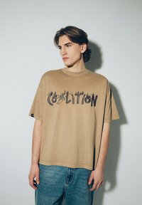 Beżowy bawełniany t-shirt z krótkimi rękawami, z czarnym napisem w stylu płomieni "Coalition". W zestawie z niebieskimi dżinsami.