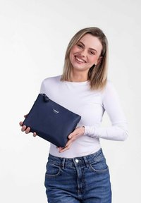 Námořnická kožená kabelka s hladkým povrchem a zipem, s logem na přední straně. Drží ji osoba oblečená v bílé košili s dlouhým rukávem.