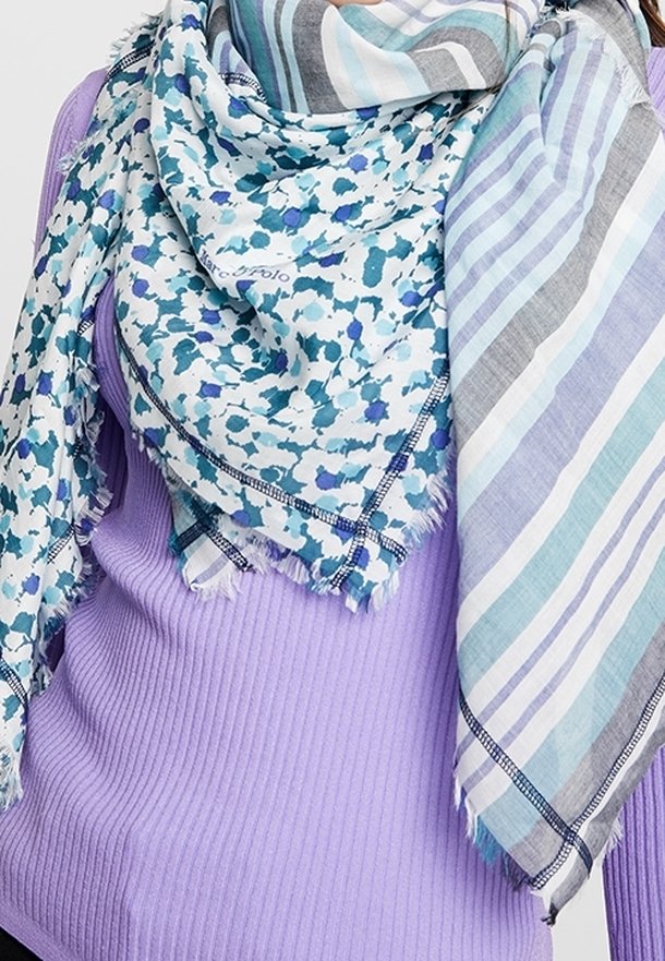 Personne portant un pull violet côtelé et deux écharpes à motifs, l'une florale et l'autre rayée, drapées autour du cou et des épaules.