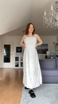 Vestido branco, sem mangas, com padrões florais bordados, design fluido e corte a meio comprimento. Usado com sapatos de plataforma pretos e meias.
