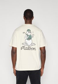 Μπεζ βαμβακερό μπλουζάκι με εικόνα ενός χιονάνθρωπου ντυμένου στα πράσινα που κρατάει μια ράβδο γκολφ, με τη λέξη «Malbon» γραμμένη κάτω από το σχέδιο.