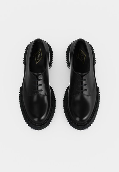 Paire de chaussures en cuir noir brillant à lacets avec semelles épaisses et dentelées, vues de dessus sur un fond clair uni.