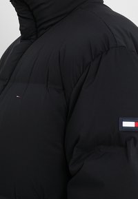 Giacca a vento nera con collo alto, caratterizzata da una toppa con logo in rosso, bianco e blu sulla manica, e una texture liscia e opaca.