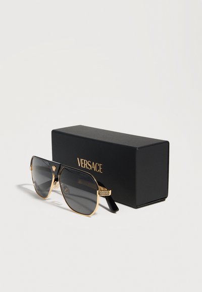 Μαύρα και χρυσά γυαλιά ηλίου Versace aviator με σκούρους φακούς δίπλα σε μια μαύρη θήκη Versace σε απλό ανοιχτόχρωμο φόντο.