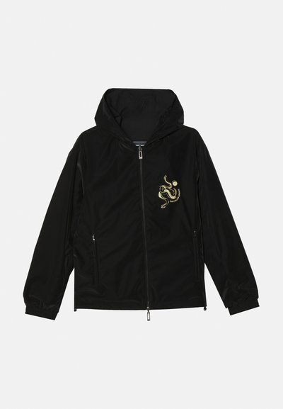 Casaco preto com fecho à frente e capuz, apresentando um design bordado a dourado no lado esquerdo do peito, punhos elásticos e dois bolsos laterais.