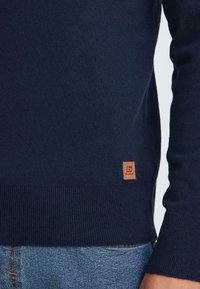 Suéter de manga larga azul marino con dobladillo acanalado y pequeña etiqueta de cuero marrón de la marca, combinado con pantalones vaqueros de denim azul.