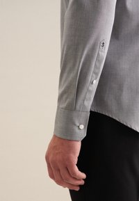 Maglietta grigia a maniche lunghe con vestibilità slim, caratterizzata da una texture liscia, polsini con bottoni e un dettaglio ricamato vicino al bottone del maniche.