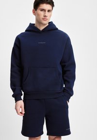 Mørkeblå hettegenser med en frontlomme og ribbestrikkede mansjetter, med en liten logo på brystet. Sammen med matchende mørkeblå shorts.