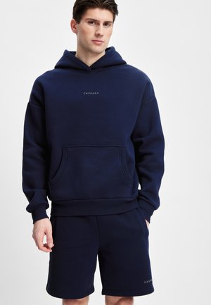Mørkeblå hettegenser med en frontlomme og ribbestrikkede mansjetter, med en liten logo på brystet. Sammen med matchende mørkeblå shorts.