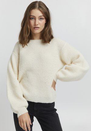 Cream chunky knit sweater with a ribbed texture, wide balloon sleeves, and a rounded neckline, paired with dark jeans.