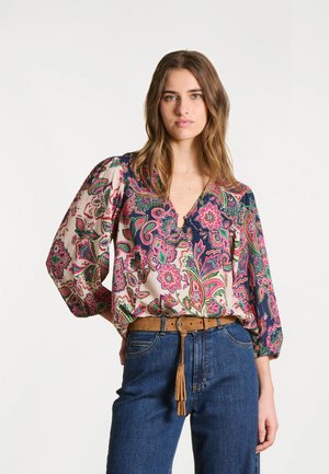 Jeune femme aux cheveux bruns lâchés portant un chemisier floral multicolore à manches bouffantes, un jean bleu et une ceinture marron à franges.